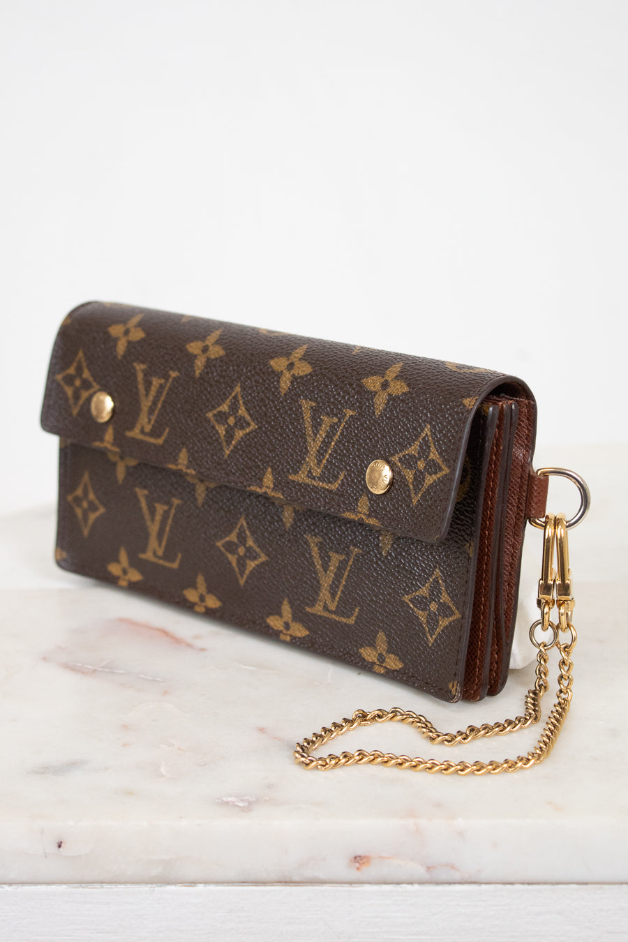 A Louis Vuitton monogram canvas clutch bag with a gold-tone chain strap, resting on a white marble surface against a white background.
