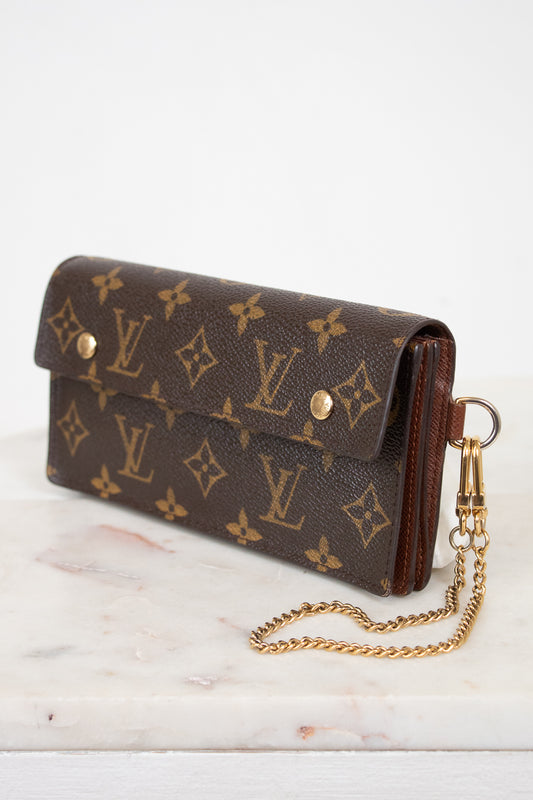A Louis Vuitton monogram canvas clutch bag with a gold-tone chain strap, resting on a white marble surface against a white background.