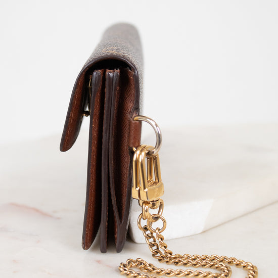 A close-up side view of a brown leather wallet or clutch with a gold-tone chain strap and clasp, resting on a white marble surface against a white background.