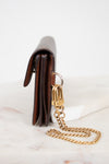 A close-up side view of a brown leather wallet or clutch with a gold-tone chain strap and clasp, resting on a white marble surface against a white background.