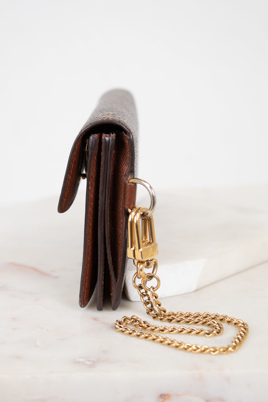 A close-up side view of a brown leather wallet or clutch with a gold-tone chain strap and clasp, resting on a white marble surface against a white background.