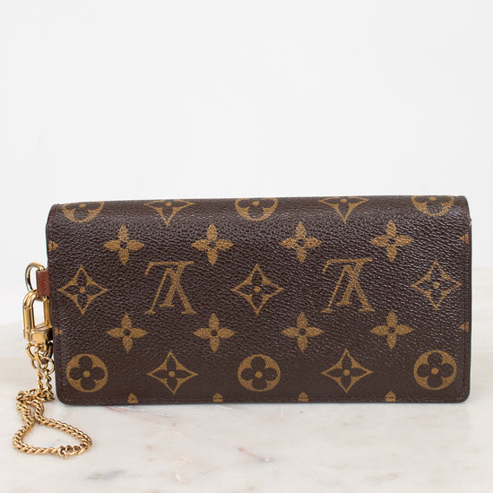 A Louis Vuitton monogram canvas wristlet clutch with a gold-tone chain strap, resting on a white marble surface against a white background.