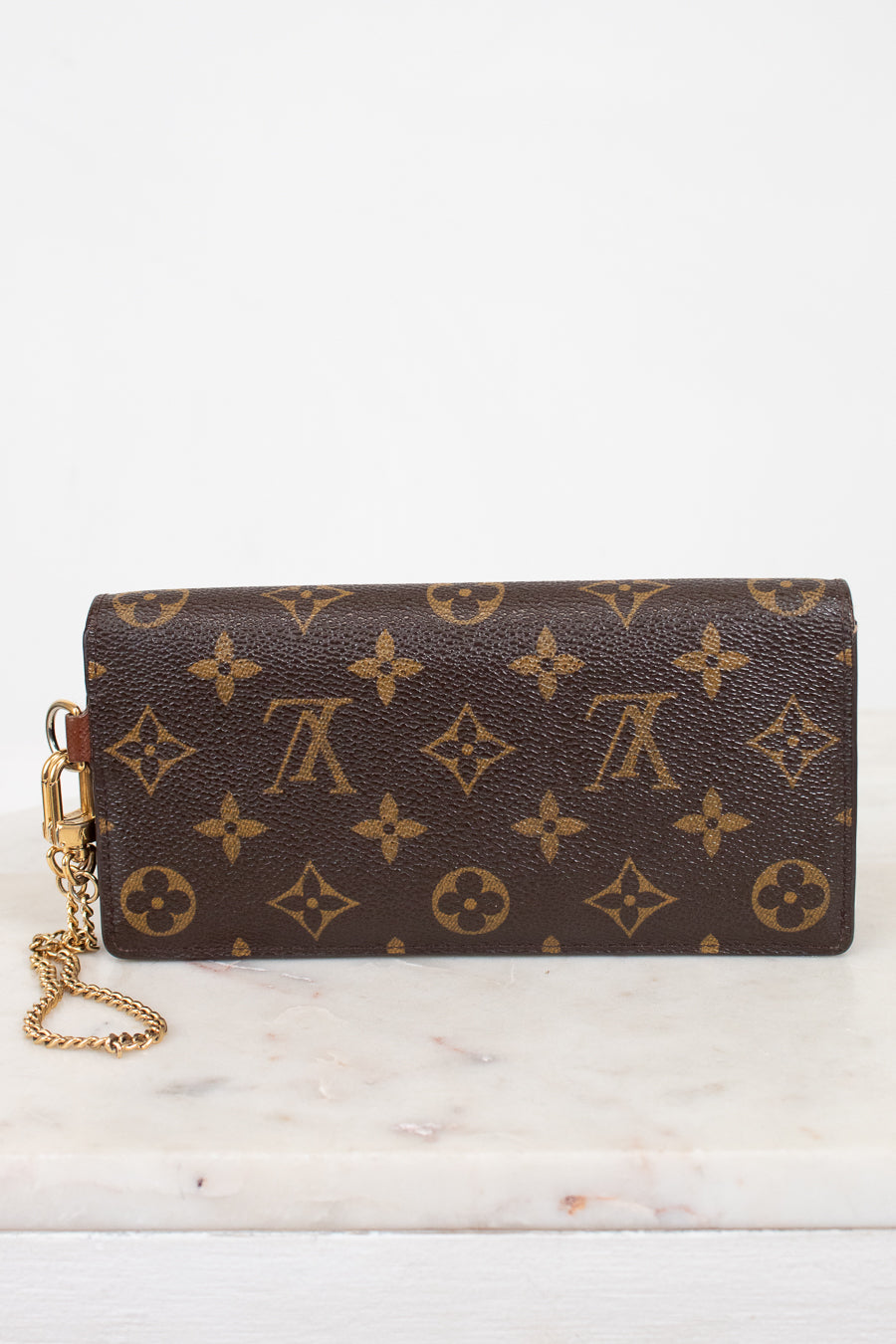 A Louis Vuitton monogram canvas wristlet clutch with a gold-tone chain strap, resting on a white marble surface against a white background.
