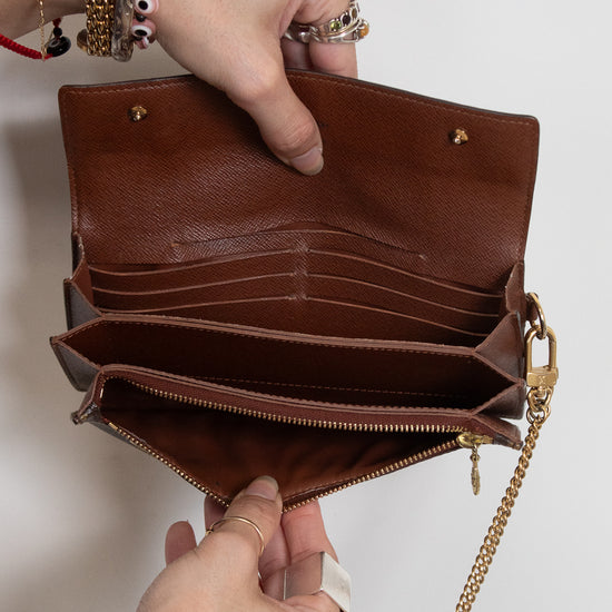 A brown leather clutch wallet shown open, held by two hands against a plain light background. The wallet features multiple card slots, a zippered compartment, and a gold-tone chain strap.