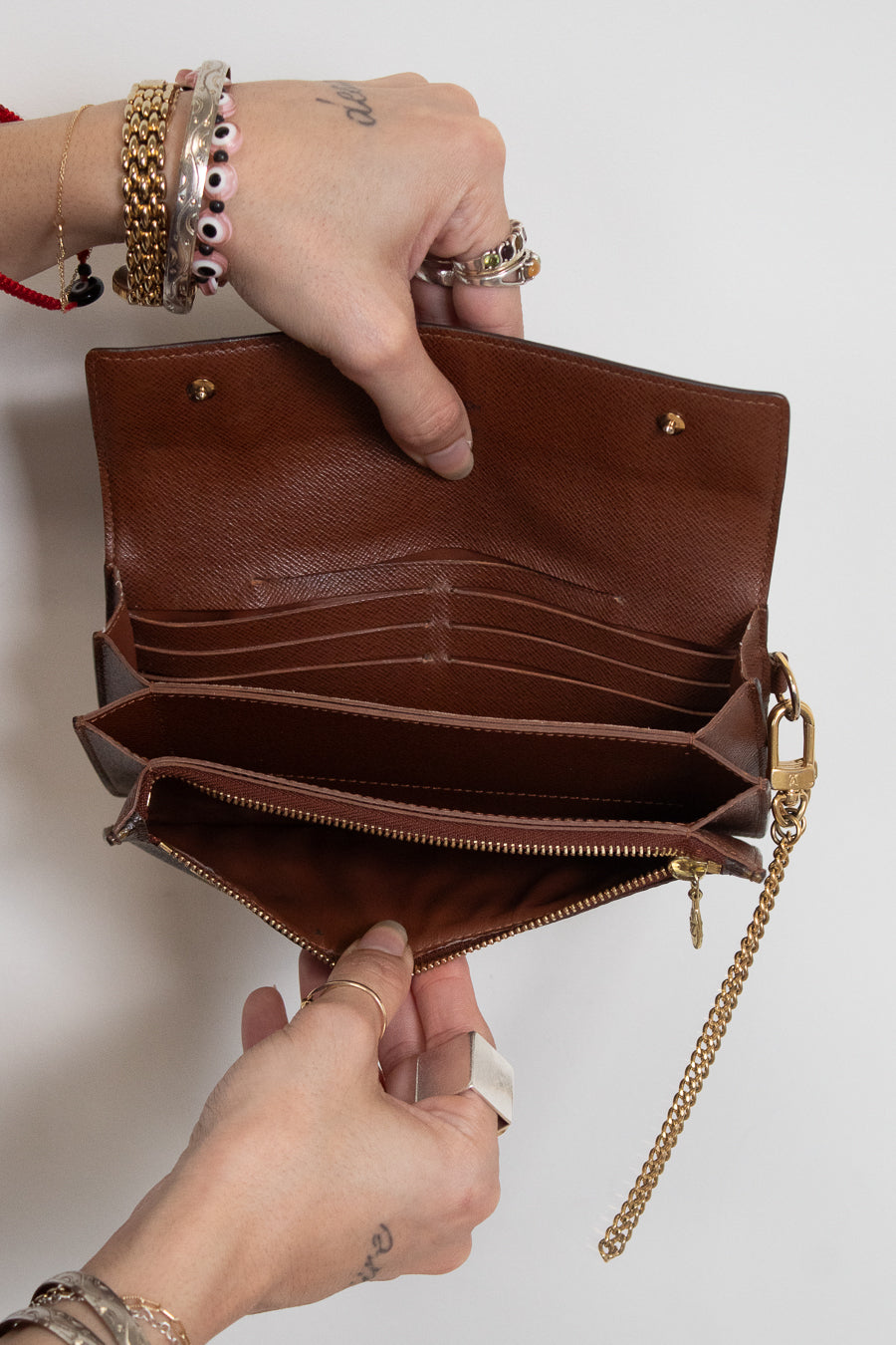 A brown leather clutch wallet shown open, held by two hands against a plain light background. The wallet features multiple card slots, a zippered compartment, and a gold-tone chain strap.