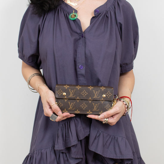 A woman wearing a dark purple puff-sleeve mini dress with a ruffled hem, holding a brown Louis Vuitton monogram clutch bag against a plain light gray background.