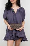 A woman wearing a dark purple puff-sleeve mini dress with a ruffled hem, holding a brown Louis Vuitton monogram clutch bag against a plain light gray background.