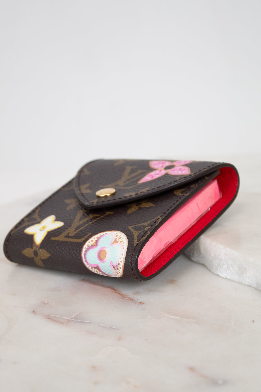A Louis Vuitton monogram post-it note holder with a bright red interior, decorated with colorful details, resting on a white marble surface.