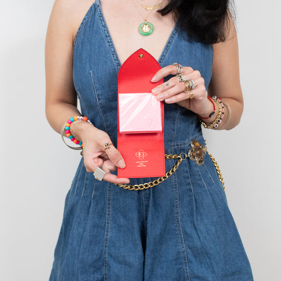A woman wearing a blue denim romper and holding a red envelope-style post-it note holder, photographed against a light gray studio background.