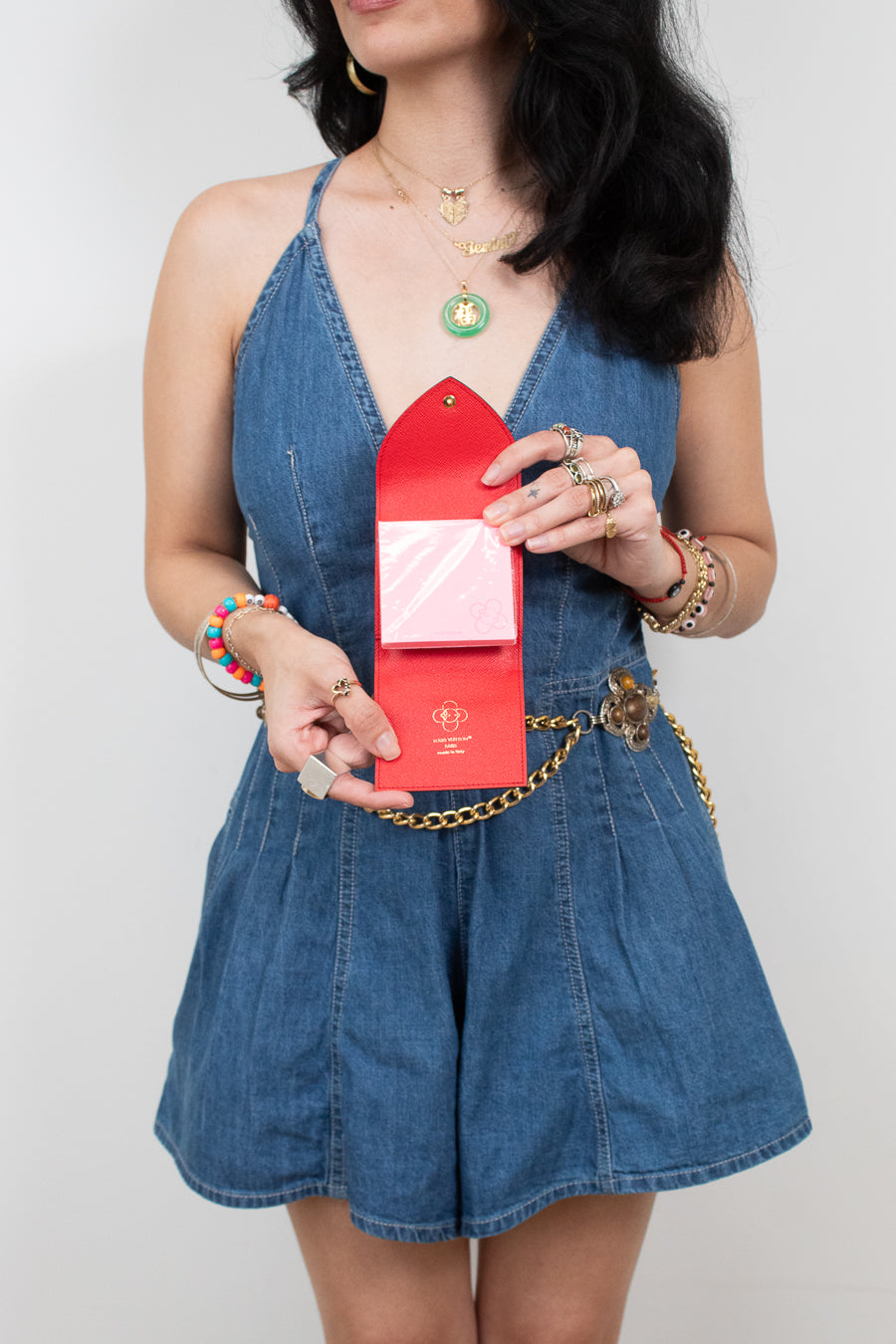 A woman wearing a blue denim romper and holding a red envelope-style post-it note holder, photographed against a light gray studio background.
