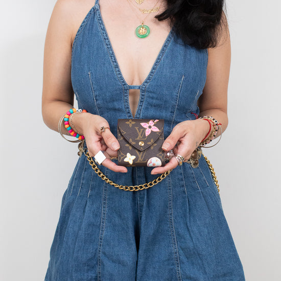 A woman wearing a blue denim romper holds a small brown monogram post-it note holder against a plain light gray background.
