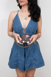 A woman wearing a blue denim romper holds a small brown monogram post-it note holder against a plain light gray background.