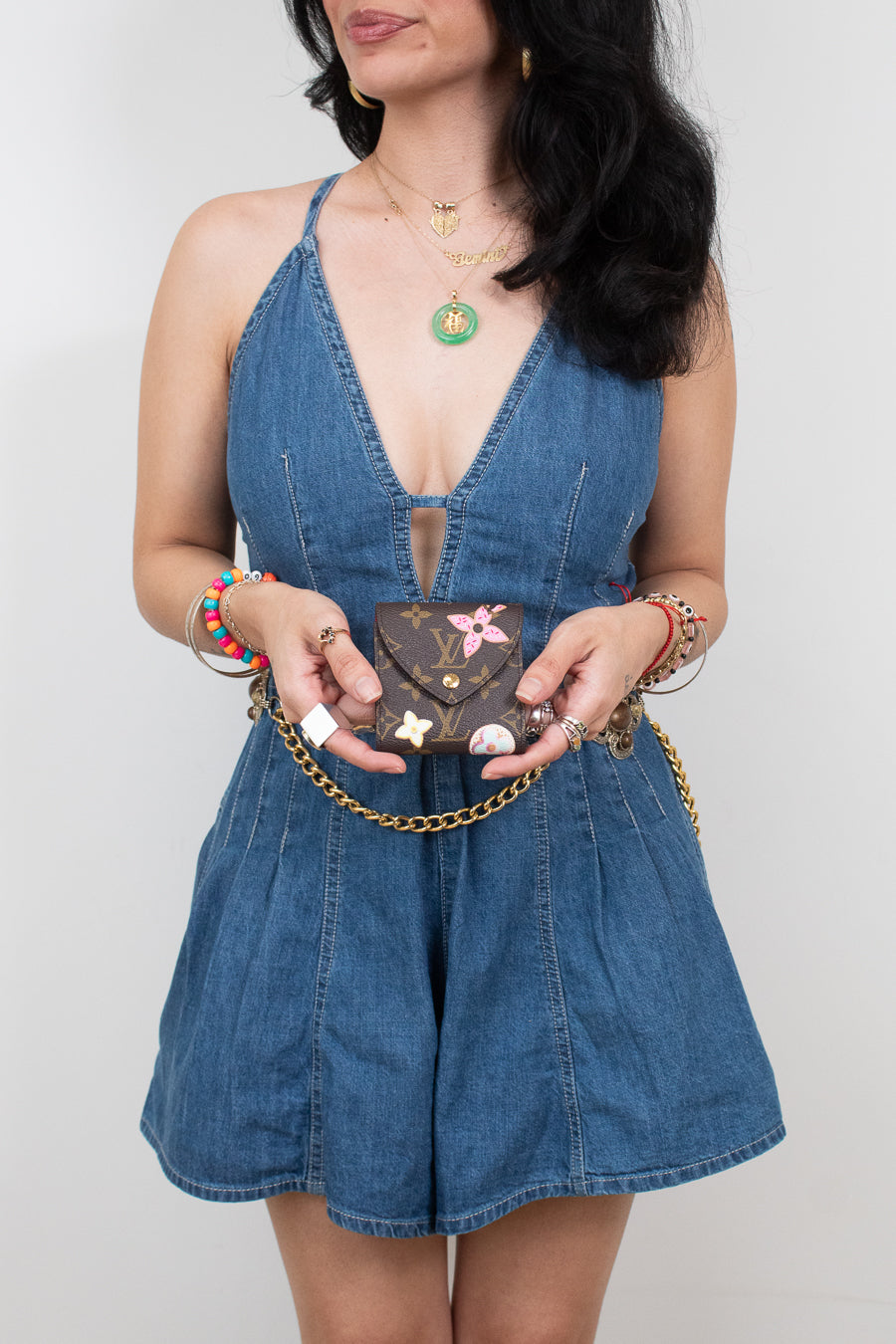 A woman wearing a blue denim romper holds a small brown monogram post-it note holder against a plain light gray background.