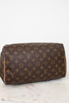 A Louis Vuitton monogram canvas shoulder bag in brown and tan, resting on a white marble surface against a white background.