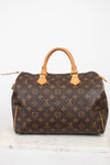 A Louis Vuitton monogram canvas Speedy handbag in dark brown with tan leather handles and trim, displayed on a white marble surface against a white background.