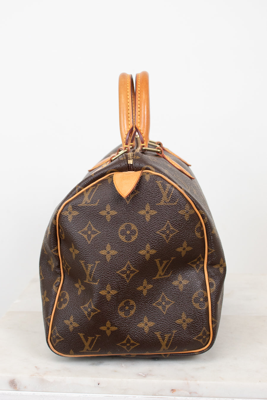 A Louis Vuitton monogram Speedy bag in brown coated canvas with tan leather trim and handles, shown from the side on a light gray background.