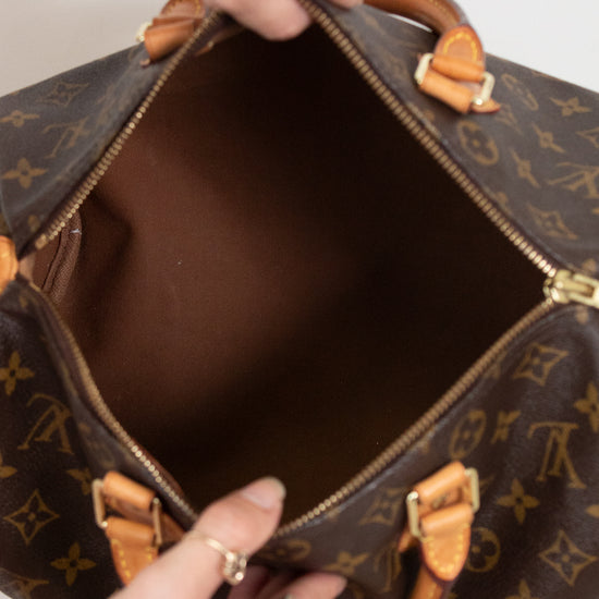 A close-up of a Louis Vuitton monogram Speedy bag being opened, showing the interior lining and gold-tone hardware, with hands holding the zipper tabs.