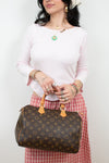 A woman wearing a light pink long-sleeve top and a red-and-white gingham skirt holds a brown Louis Vuitton monogram Speedy handbag with tan leather handles against a plain white background.