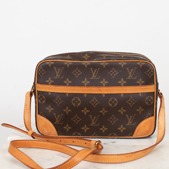A Louis Vuitton monogram coated canvas crossbody camera bag with tan leather trim and strap, displayed on a white marble surface against a white background.