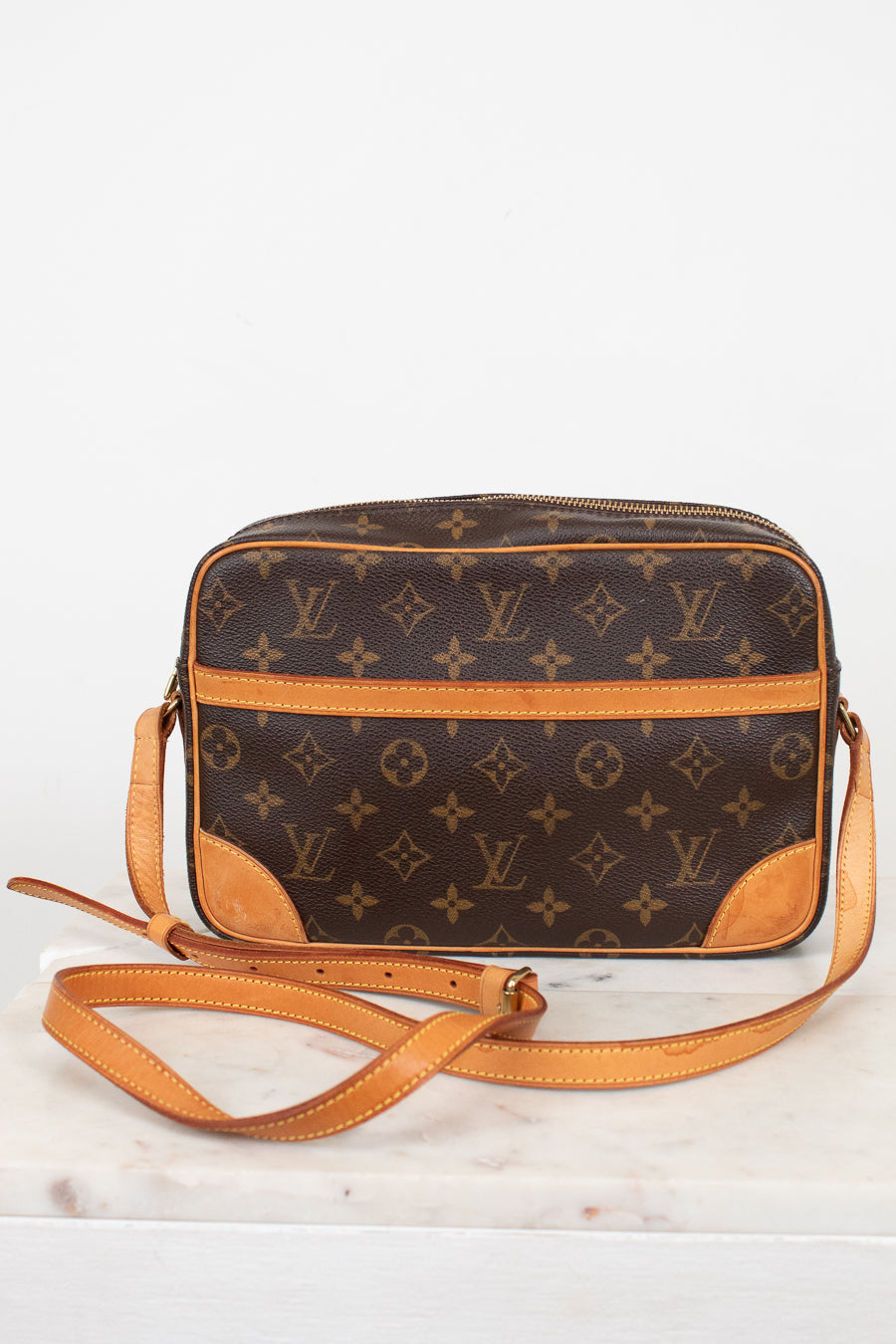 A Louis Vuitton monogram coated canvas crossbody camera bag with tan leather trim and strap, displayed on a white marble surface against a white background.