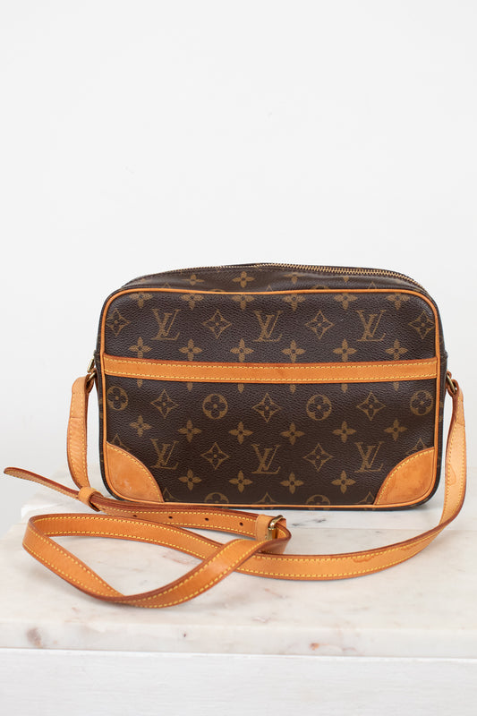 A Louis Vuitton monogram coated canvas crossbody camera bag with tan leather trim and strap, displayed on a white marble surface against a white background.