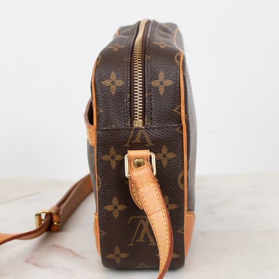 A Louis Vuitton monogram coated canvas crossbody bag shown from the side profile, featuring the classic brown and tan monogram pattern with a gold-tone zipper and leather strap on a light marble surface.