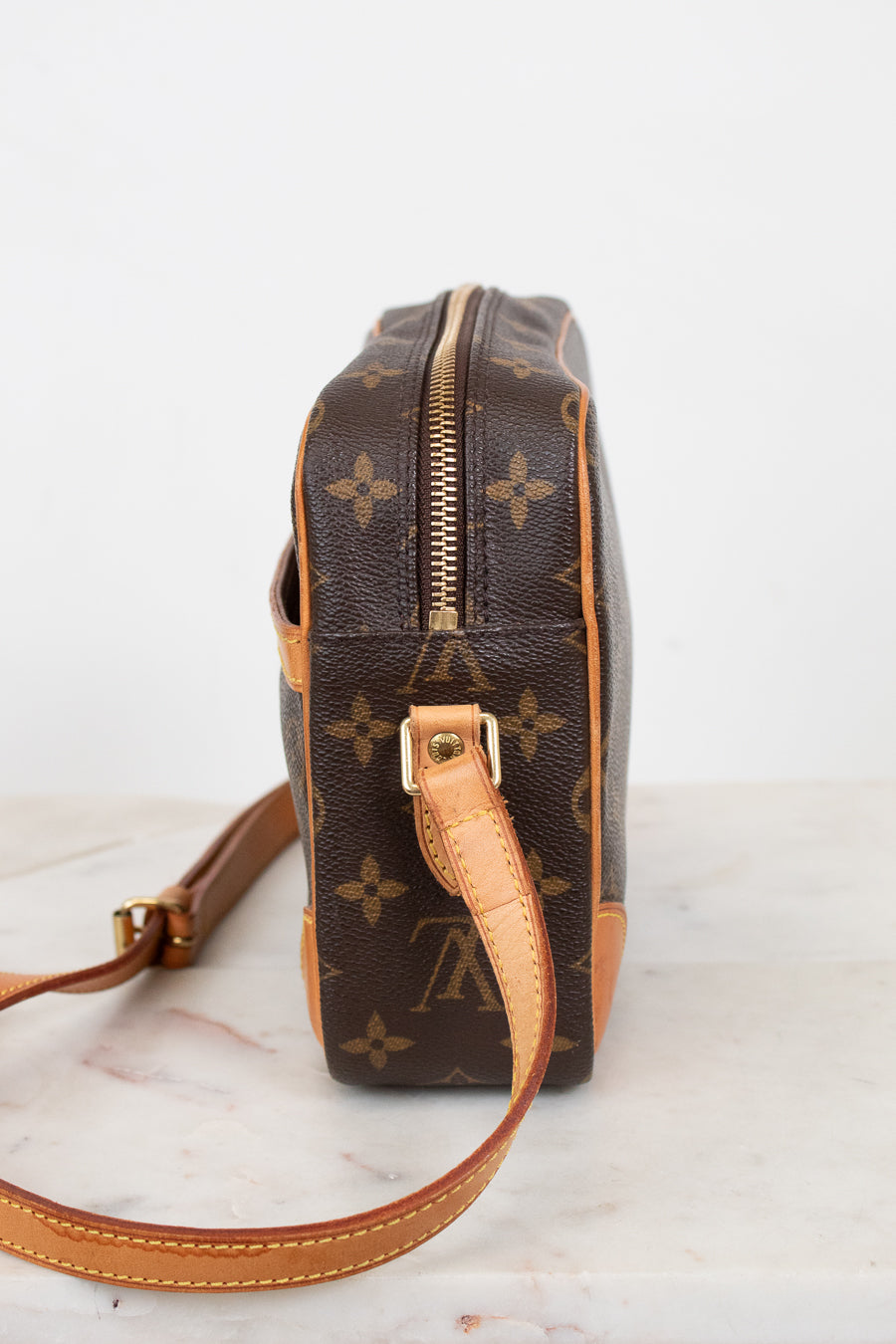 A Louis Vuitton monogram coated canvas crossbody bag shown from the side profile, featuring the classic brown and tan monogram pattern with a gold-tone zipper and leather strap on a light marble surface.