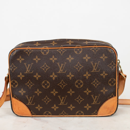 A Louis Vuitton monogram canvas crossbody bag with tan leather trim and corner details, resting on a white marble surface against a white background.