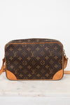 A Louis Vuitton monogram canvas crossbody bag with tan leather trim and corner details, resting on a white marble surface against a white background.