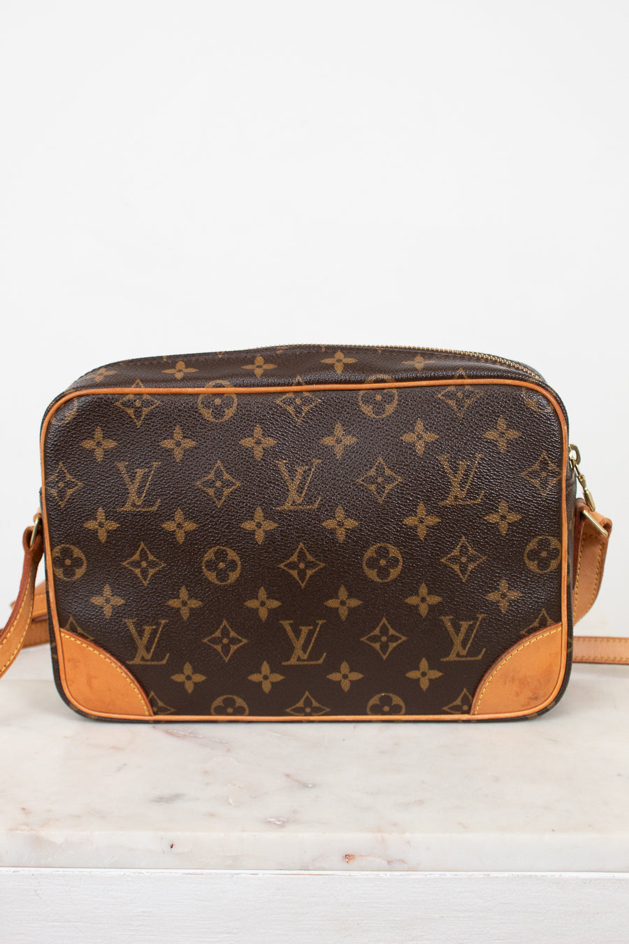 A Louis Vuitton monogram canvas crossbody bag with tan leather trim and corner details, resting on a white marble surface against a white background.