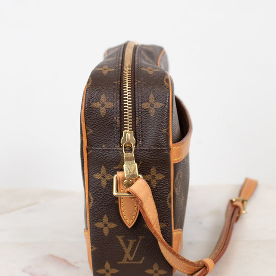 A Louis Vuitton monogram coated canvas camera bag shown from the side profile, featuring the classic brown and tan monogram pattern with gold-tone hardware and a leather shoulder strap resting on a light marble surface against a white background.