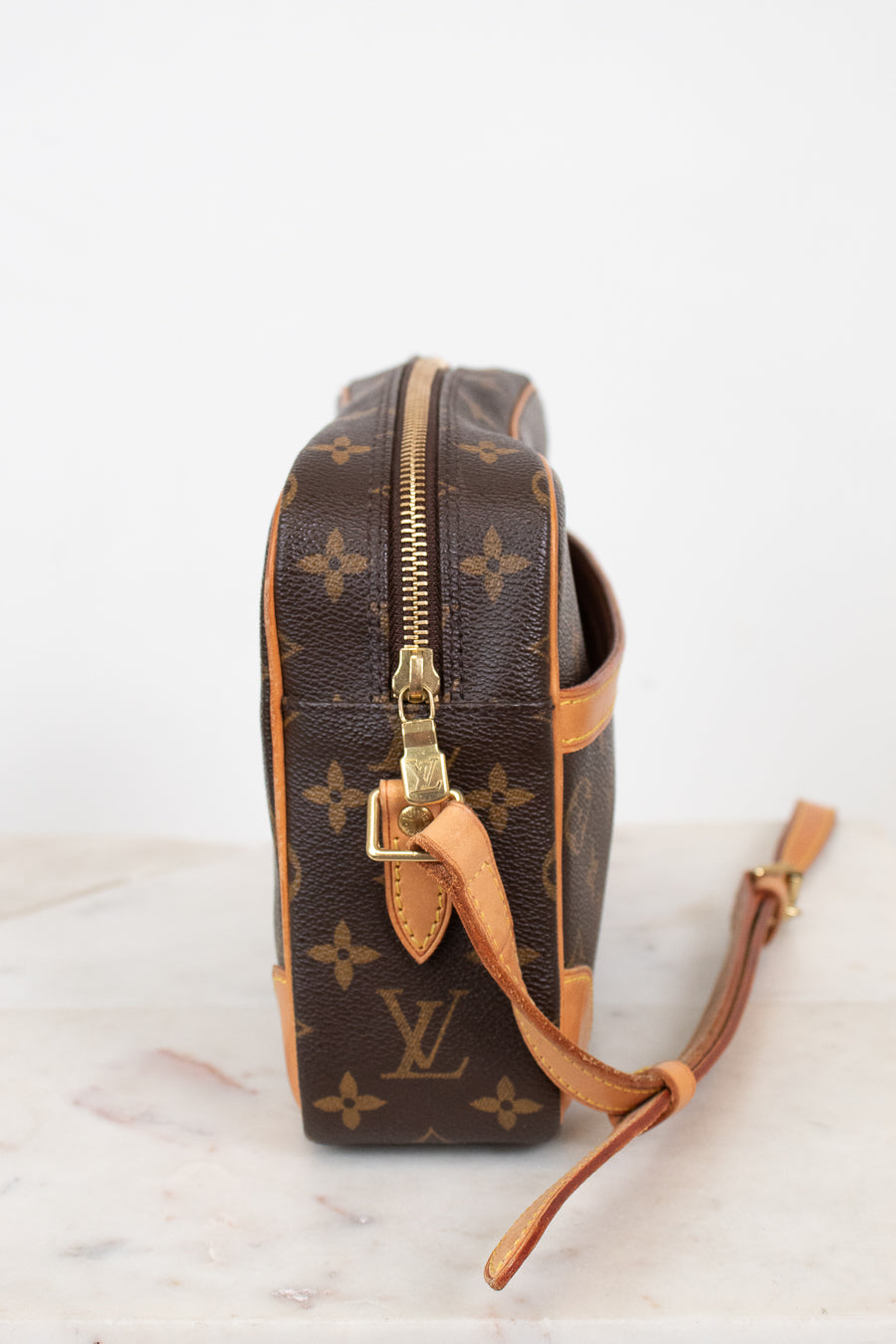 A Louis Vuitton monogram coated canvas camera bag shown from the side profile, featuring the classic brown and tan monogram pattern with gold-tone hardware and a leather shoulder strap resting on a light marble surface against a white background.