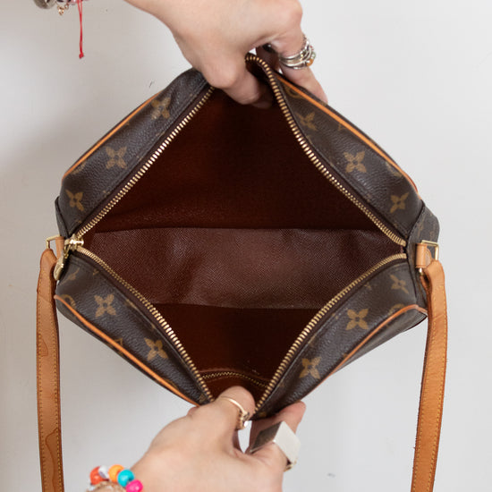 A Louis Vuitton shoulder bag in brown monogram canvas is shown open, with a person's hands holding it to display the interior. The bag features gold-tone hardware, a brown leather interior, and tan leather shoulder straps against a plain white background.