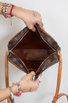 A Louis Vuitton shoulder bag in brown monogram canvas is shown open, with a person's hands holding it to display the interior. The bag features gold-tone hardware, a brown leather interior, and tan leather shoulder straps against a plain white background.