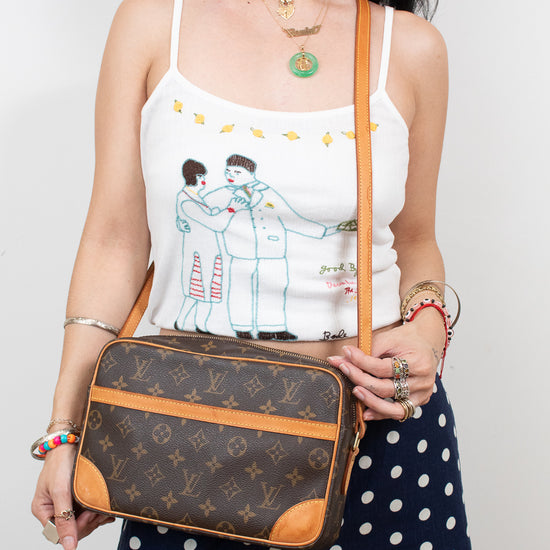A woman wearing a white spaghetti-strap tank top with a cartoon illustration and navy blue polka-dot pants, holding a Louis Vuitton monogram crossbody bag with tan leather trim.