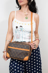 A woman wearing a white spaghetti-strap tank top with a cartoon illustration and navy blue polka-dot pants, holding a Louis Vuitton monogram crossbody bag with tan leather trim.
