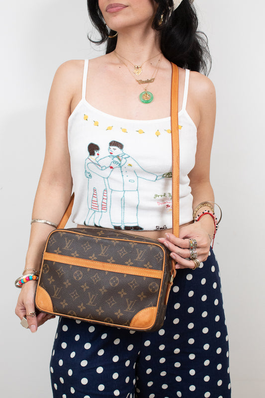A woman wearing a white spaghetti-strap tank top with a cartoon illustration and navy blue polka-dot pants, holding a Louis Vuitton monogram crossbody bag with tan leather trim.