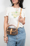Person wearing a white t-shirt with a graphic design, blue jeans, and holding a brown handbag.