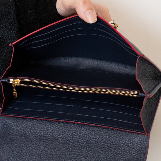 An open black pebbled leather clutch or wallet with red piping, gold-tone hardware, and a hand holding it open. A gold-tone V logo is visible on the lower front.
