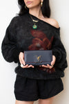 A model wearing a fuzzy black off-the-shoulder sweater and black shorts holds a dark navy blue textured leather clutch with a gold-tone Louis Vuitton monogram logo.