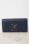 A Louis Vuitton pebbled leather long wallet in navy blue with a gold-tone LV logo clasp, resting on a light marble surface against a white background.