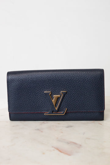 A Louis Vuitton pebbled leather long wallet in navy blue with a gold-tone LV logo clasp, resting on a light marble surface against a white background.