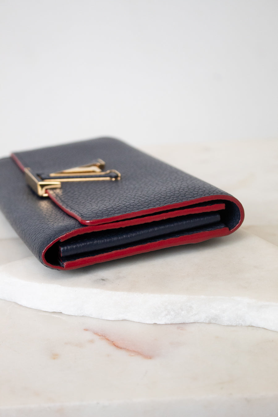 A black pebbled leather wallet with a gold-tone metal clasp and a contrasting red interior lining, resting on a white marble slab against a light gray background.