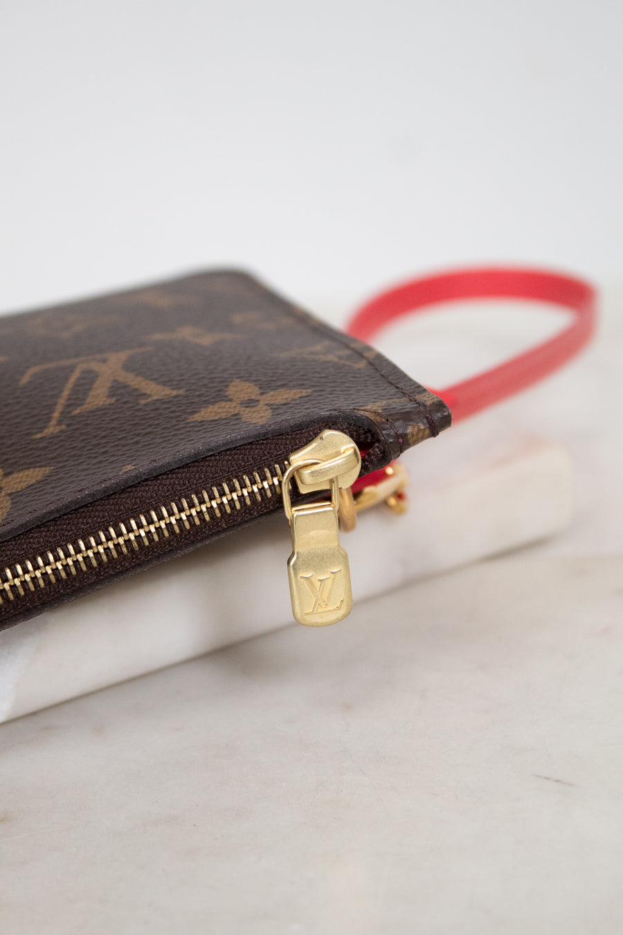A close-up of a Louis Vuitton monogram canvas zip pouch with a gold-tone zipper pull and a bright red wristlet strap, resting on a white marble surface.