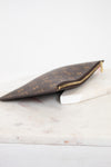 A Louis Vuitton monogram canvas zip pouch in brown and gold, resting on a white marble block against a white background.