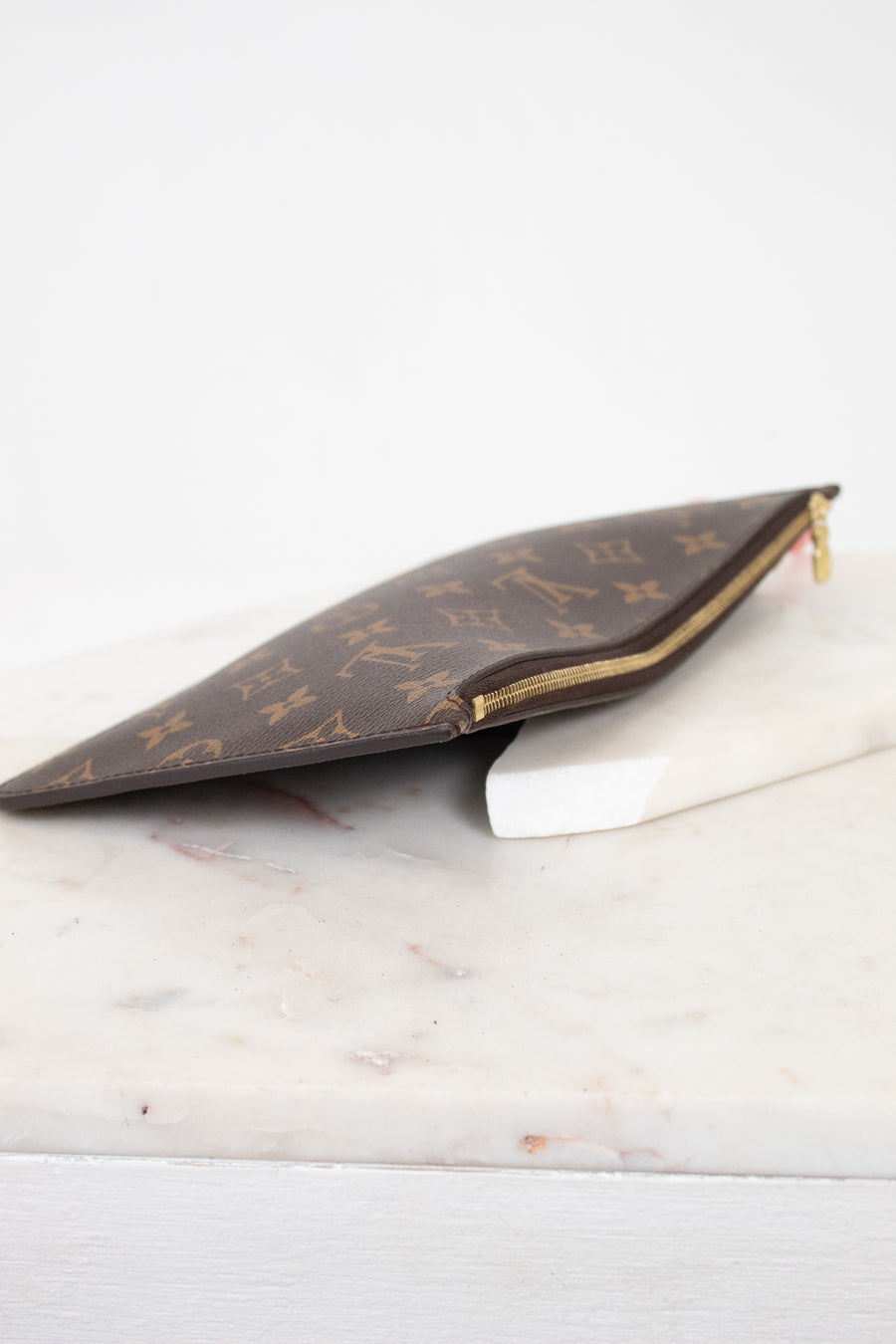 A Louis Vuitton monogram canvas zip pouch in brown and gold, resting on a white marble block against a white background.