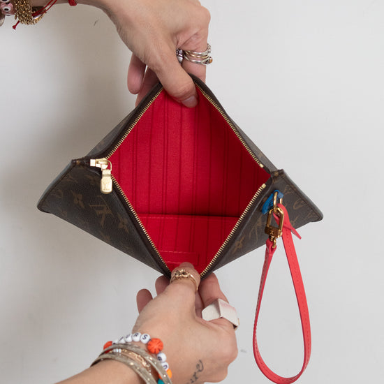 A Louis Vuitton monogram clutch pouch with a bright red interior lining and a red wristlet strap, held open by two hands wearing various bracelets and rings against a plain white background.