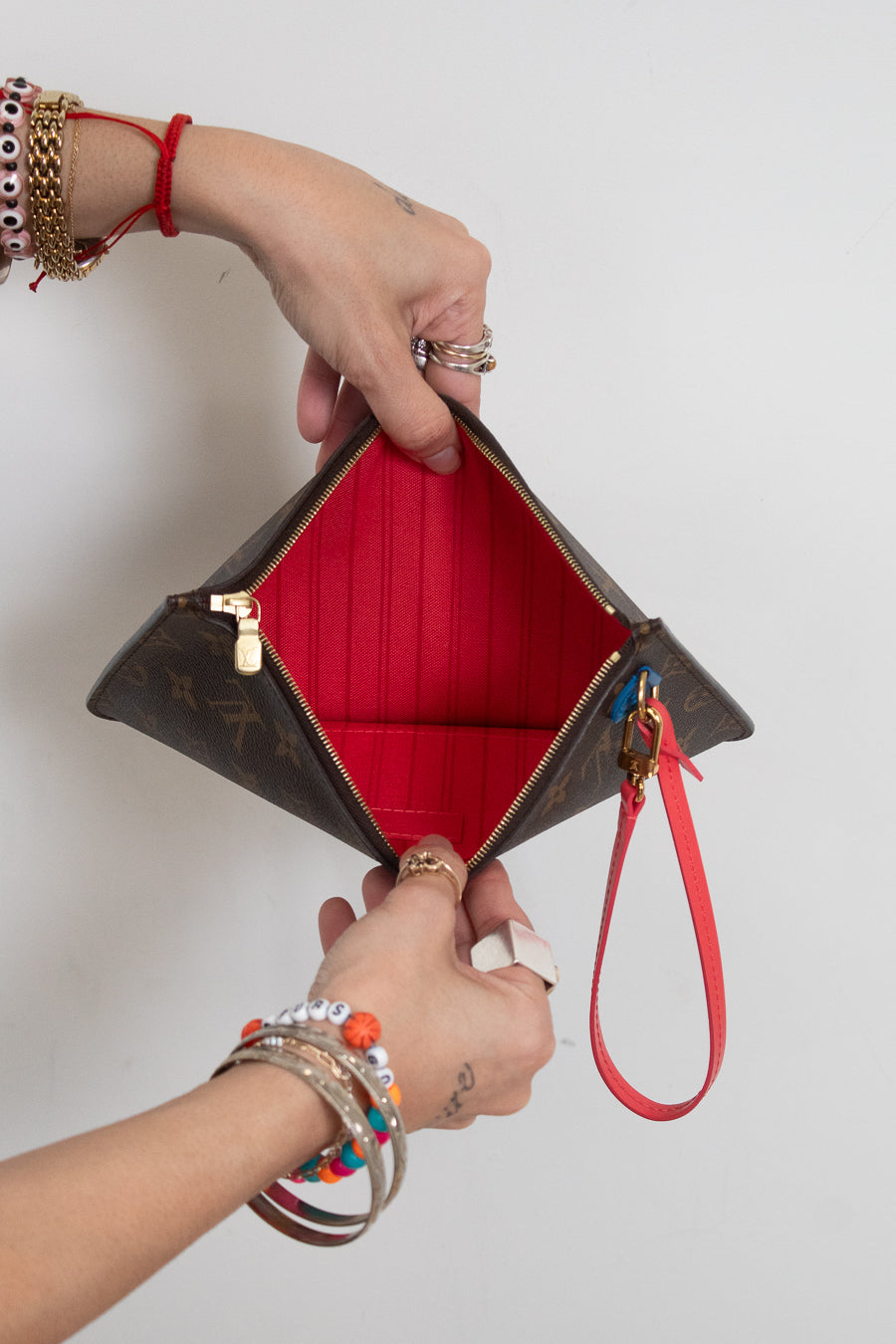 A Louis Vuitton monogram clutch pouch with a bright red interior lining and a red wristlet strap, held open by two hands wearing various bracelets and rings against a plain white background.