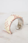 A Louis Vuitton cuff bracelet featuring cream-colored monogram canvas with a light pink border and silver-tone metal spikes, resting on a light marble surface.