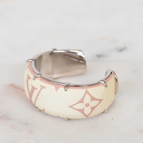 A Louis Vuitton cuff bracelet in beige monogram canvas with silver-tone metal studs and a silver interior, resting on a light marble surface.